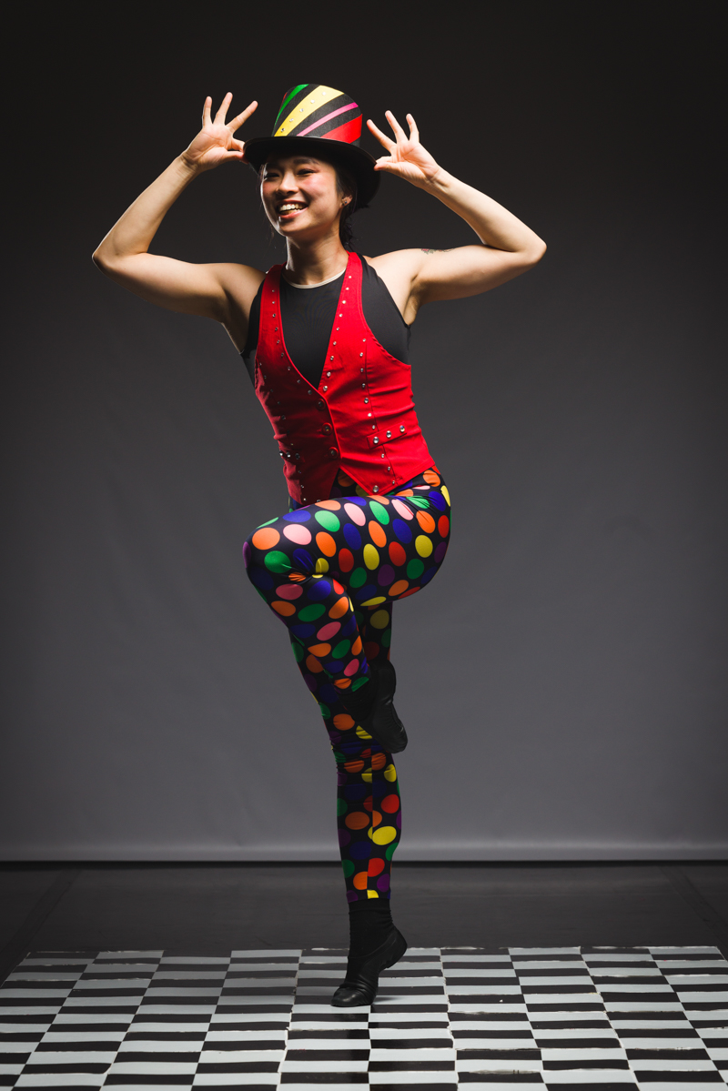 2023-05-13Ormao(285of357)_1 Nicole in a black leotard, bejeweled red vest, black top hat with colored ribbon and rhinestones, and rainbow polka dot leggings in a high parallel retire pose with both hands touching the hat